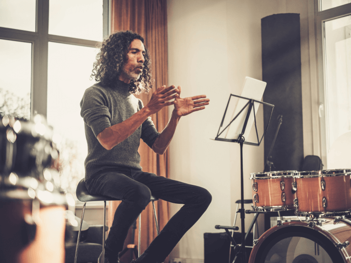 A teacher sits at the front of the classroom, gesturing forwards in a deliberate way. A music stand is in front of him, and a drum kit is also in shot.
