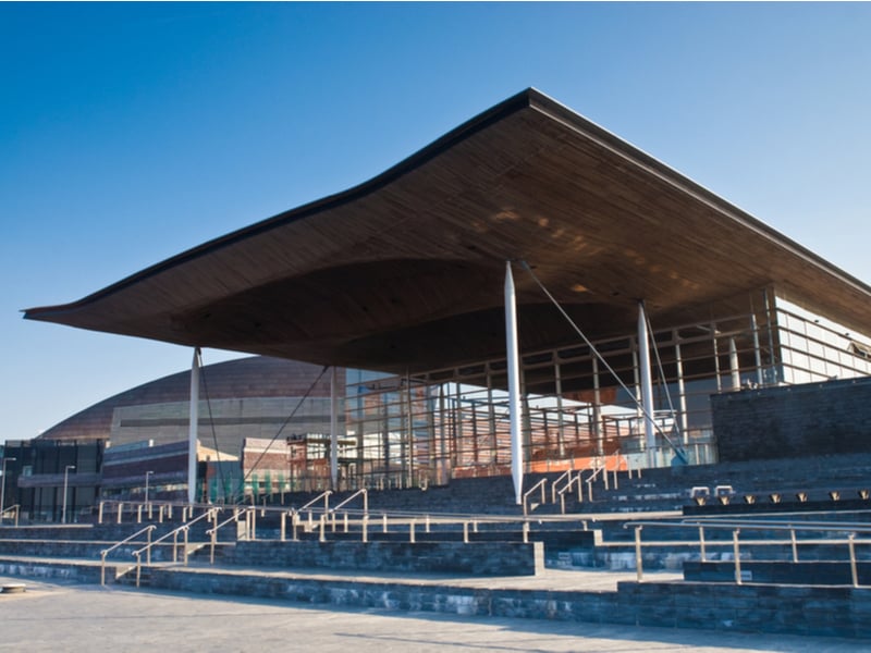 The Welsh Parliament building at the Cardiff Bay development, the sun is shining in a blue sky behind a very modern piece of architecture with a glass wall and a large wooden roof.