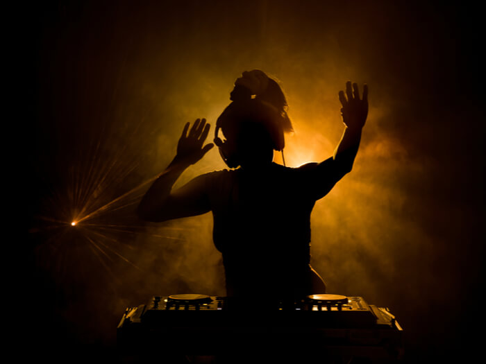 A woman is outlined against a golden yellow light standing at a DJ desk.