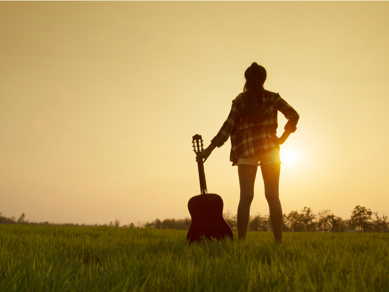 Photograph of a woman in a plaid shirt, standing in a field and looking out to a sunset.