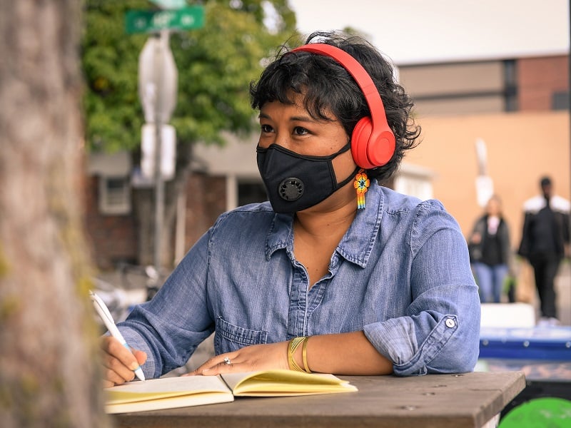 A woman sat outside is writing into a notebook, she is wearing earphones and a facemask and looking into the distance.