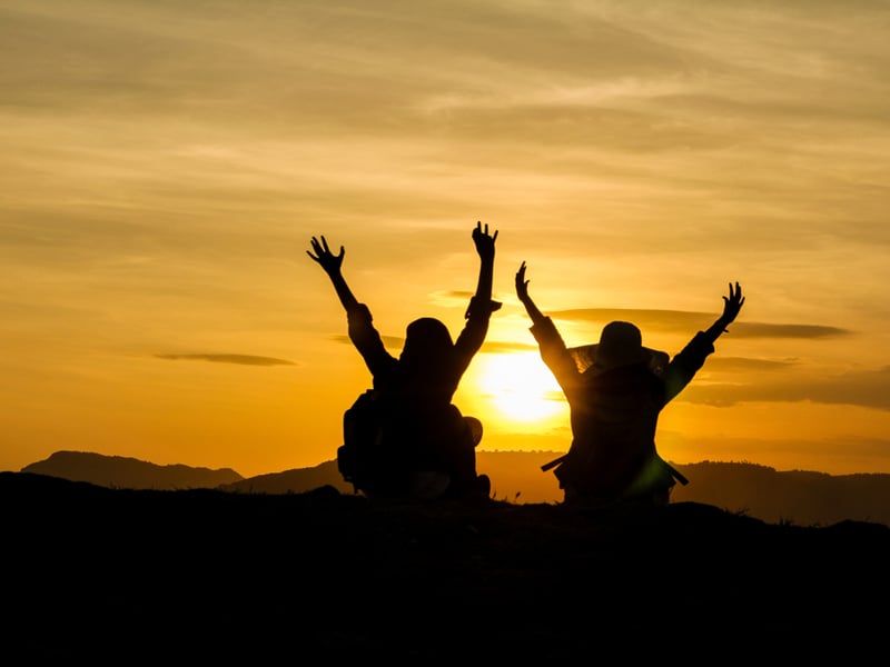 Two women sat in the mountains, raise their arms into the air as the sun rises in front of them.