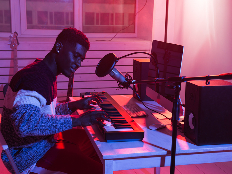 A young musician is working on a keyboard with a microphone, speakers and computer set up in front of them. There is warm but dark lighting.