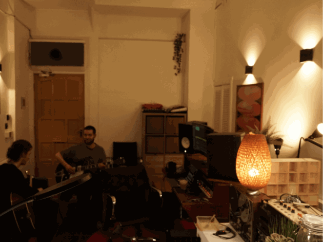 Inside of the studio, a warmly lit space with plants and lamps. Two musicians are playing guitar.